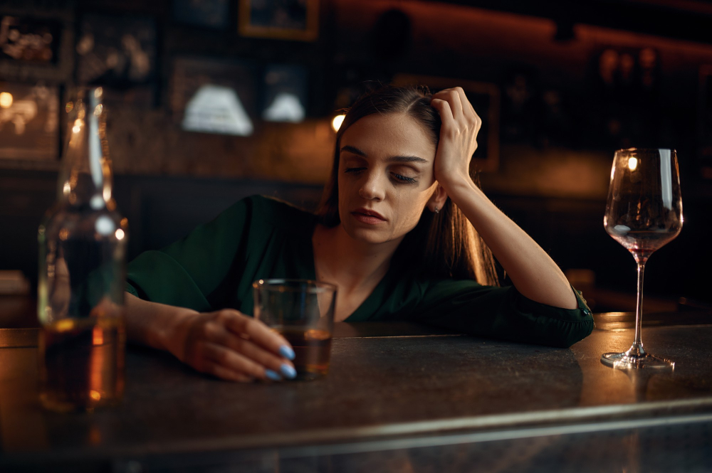 Pijana kobieta z półprzymkniętymi oczyma, podpierająca się ręką o drewniany bar w pubie i trzymająca szklankę z alkoholem