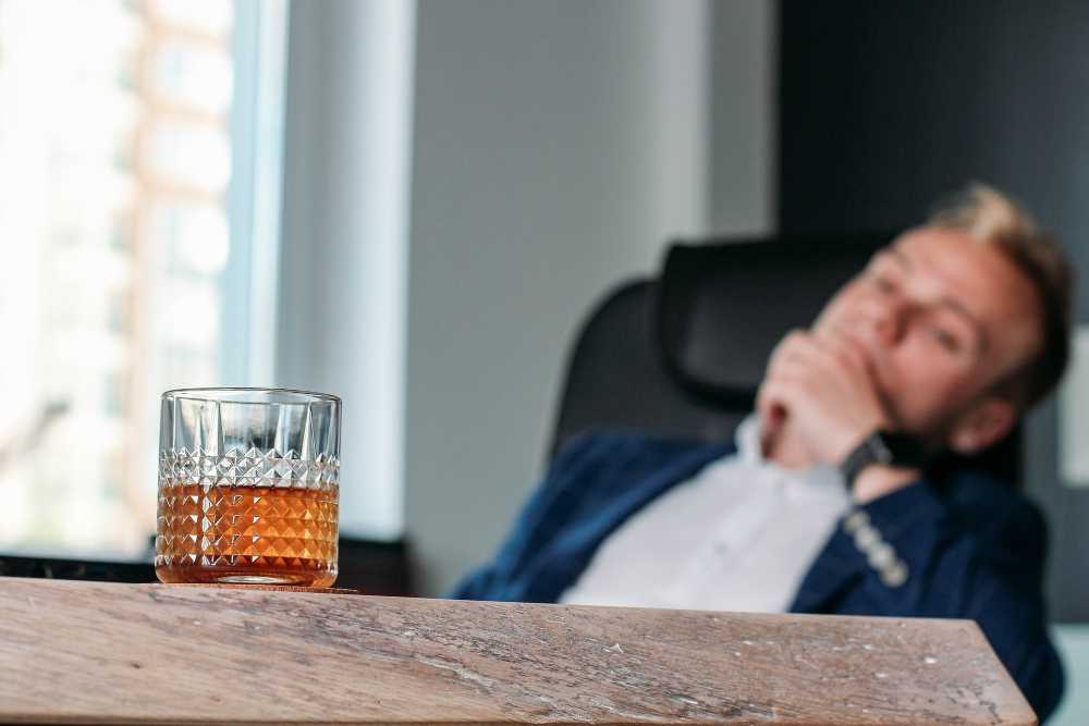 Mężczyzna siedzący na skórzanym fotelu w biurze, przed szklanką whiskey leżącej na drewnianym stole