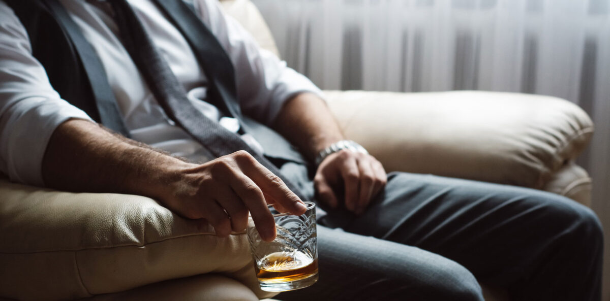 Wysokofunkcjonujący alkoholik wiedzący w fotelu w koszuli pod krawatem i eleganckiej kamizelce ze szklanką whiskey w ręku